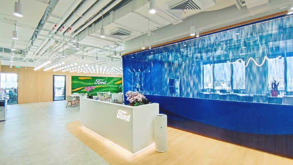 مكتب استقبال حديث في مقر فورد، يضم مكتبًا أبيض مضيئًا، وجدران زخرفية باللونين الأخضر والأزرق تحمل شعار فورد، وسقف مكشوف البنية، ومساحة عمل مفتوحة تحت إضاءة معلقة ساطعة.