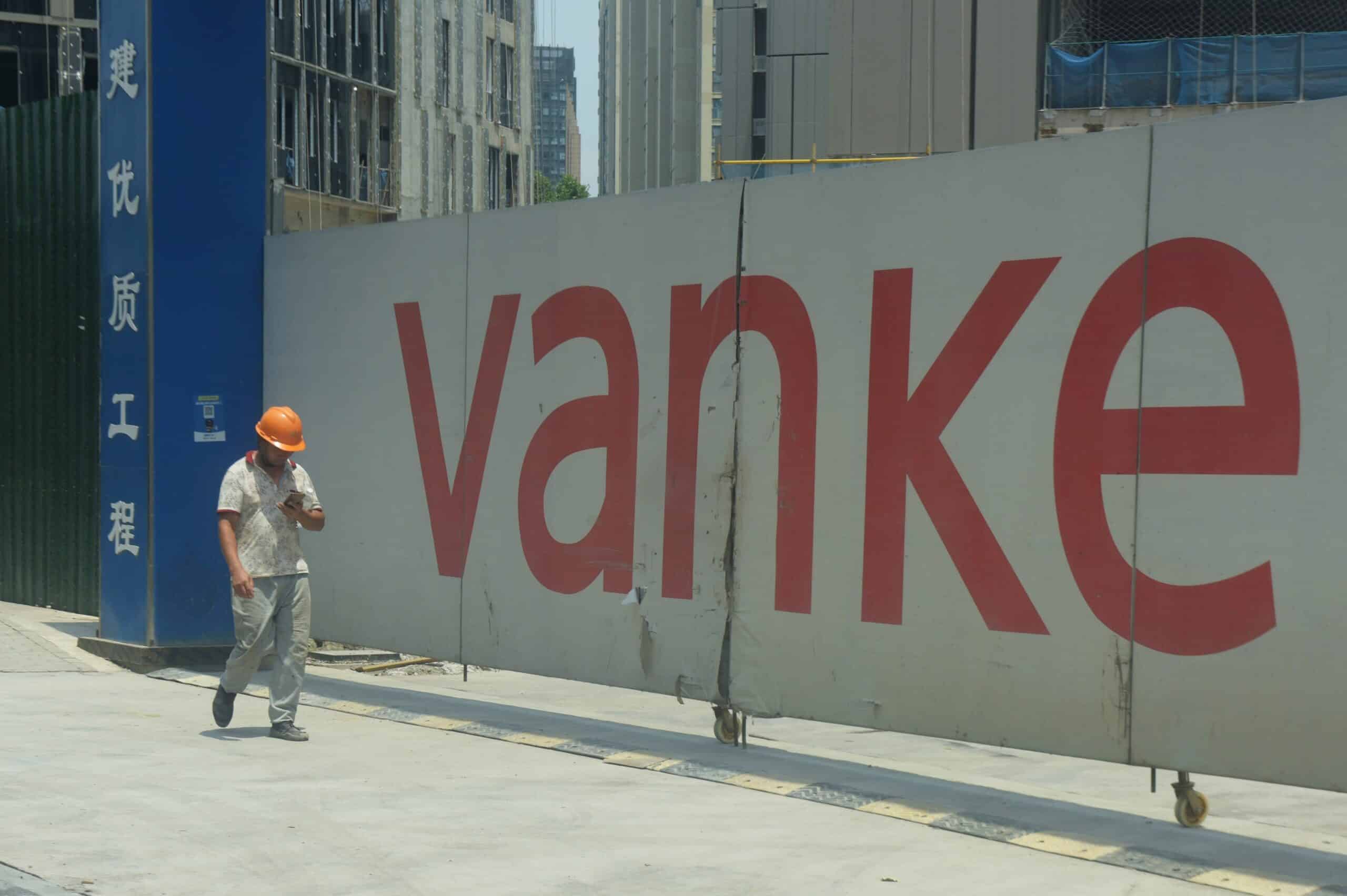A construction worker wearing an orange hard hat and light-colored clothing walks on a sidewalk in front of a large, temporary construction hoarding. The hoarding is mostly white with the large, prominent red text "vanke" written across it, and a blue panel with Chinese characters is visible on the left. The background shows parts of a tall building under construction