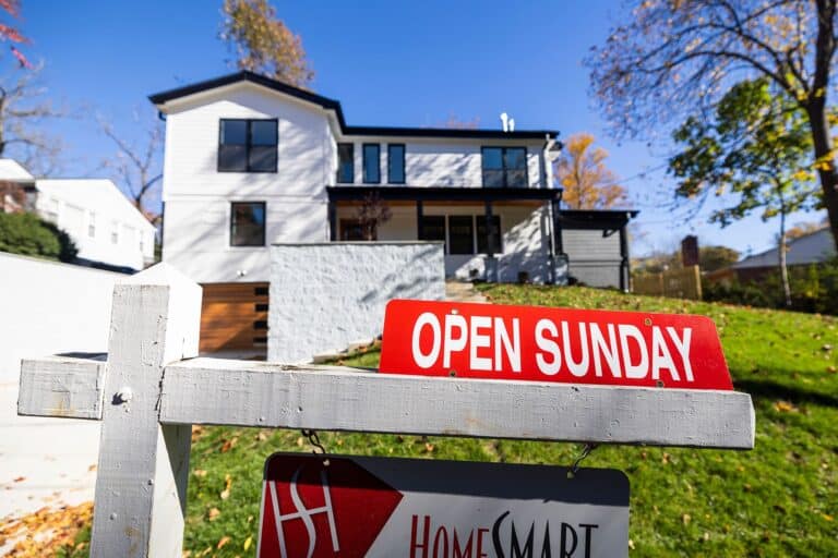 A modern white two-story house with black trim and a wooden garage door. In the foreground, a white real estate post features a bright red "Open Sunday" sign and a HomeSmart logo hanging below.