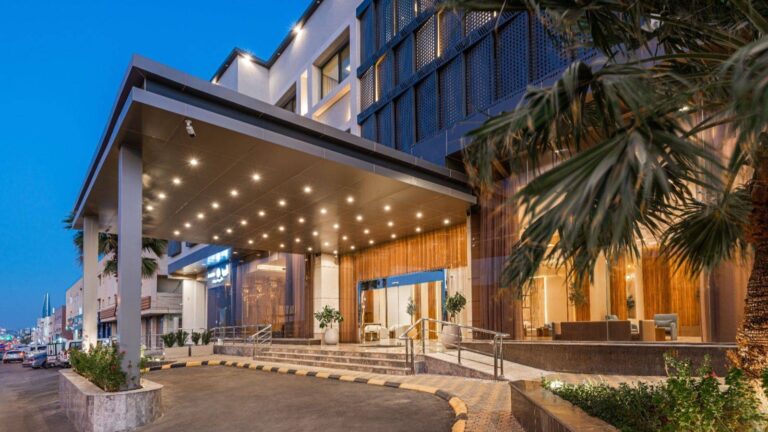 A wide-angle view of a modern hotel entrance featuring a large gray canopy with bright recessed lights, wooden wall panels, glass doors, and a palm tree in the foreground during evening.