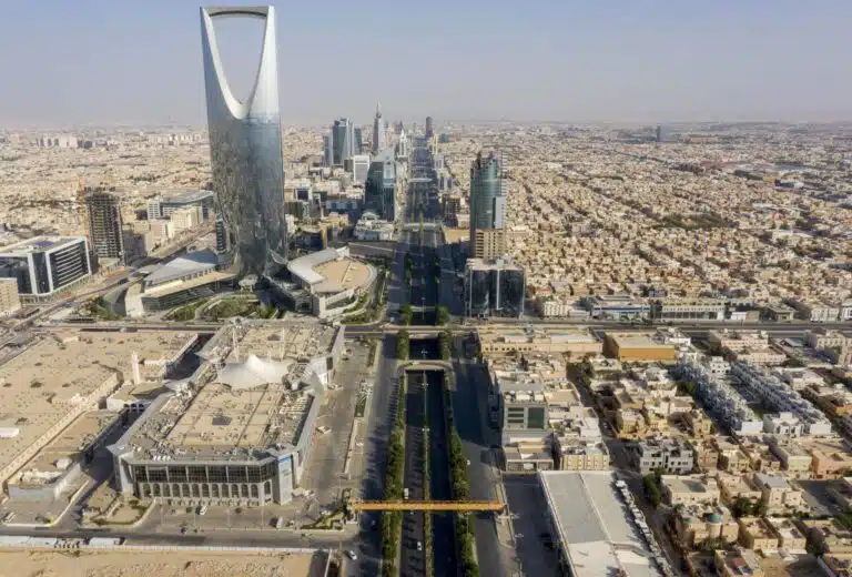 An aerial wide shot of the Riyadh skyline featuring the iconic Kingdom Centre tower and the long King Fahd Road stretching into the distance under a clear sky.