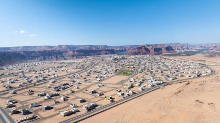 An aerial drone shot of a modern residential neighborhood in a desert valley, featuring grid-patterned streets and light-colored square houses, surrounded by vast sand and red rock mountains under a clear blue sky.