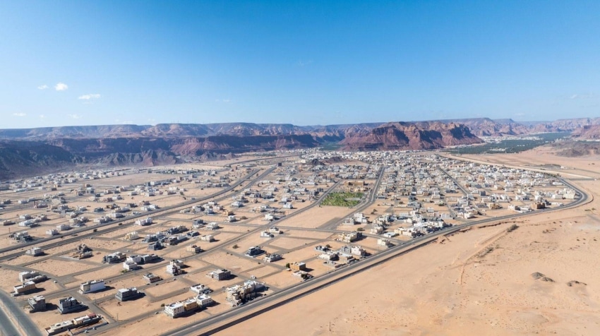 An aerial drone shot of a modern residential neighborhood in a desert valley, featuring grid-patterned streets and light-colored square houses, surrounded by vast sand and red rock mountains under a clear blue sky.