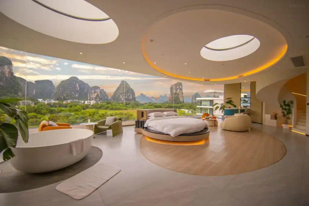 A modern hotel suite at LS Hotel in Guilin featuring a circular bed, freestanding bathtub, and floor to ceiling windows that frame the karst mountain landscape under a curved ceiling with recessed lighting. Avatar like views