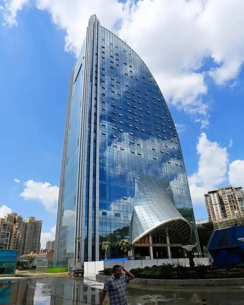 منظور من الأسفل لبرج ليبيان الدولي في قوييانغ، الصين، يُظهر واجهته الزجاجية المنحنية وهي تعكس السماء والغيوم. يضم المدخل سقفًا نحتيًا، مع أرضية رطبة تشير إلى نشاط مائي حديث.