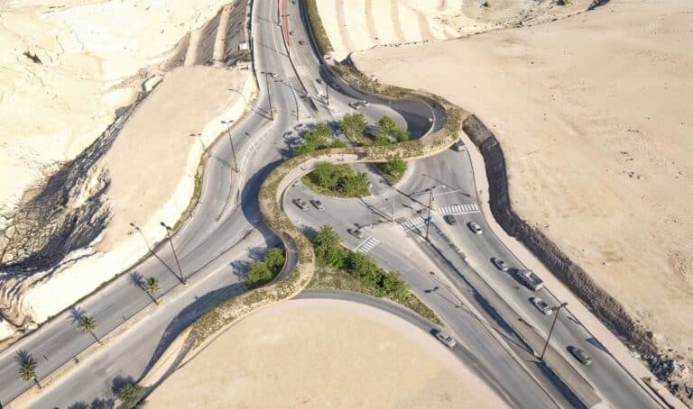 Aerial view of a serpentine road intersection in an arid landscape, with curved asphalt lanes, sparse greenery, and vehicles navigating the bends under bright daylight.