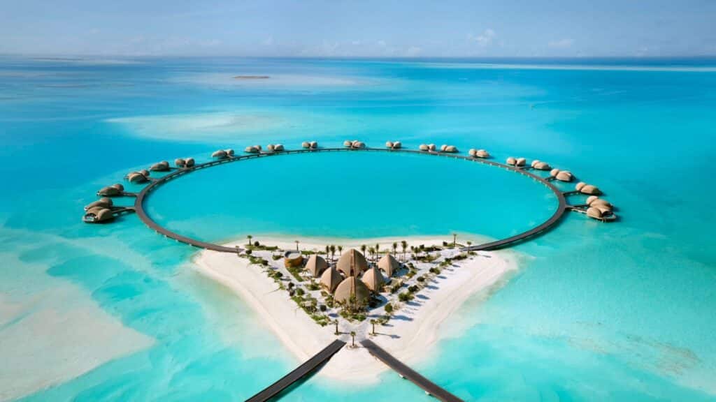 Aerial view of a circular overwater resort complex with domed villas arranged around a central lagoon, connected by walkways over turquoise ocean. Red Sea destination