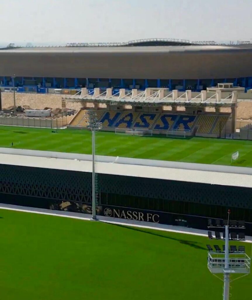 Aerial view of Al-Nassr FC&rsquo;s training pitch with adjacent stadium in background, featuring bold blue and yellow seating spelling NASSR .