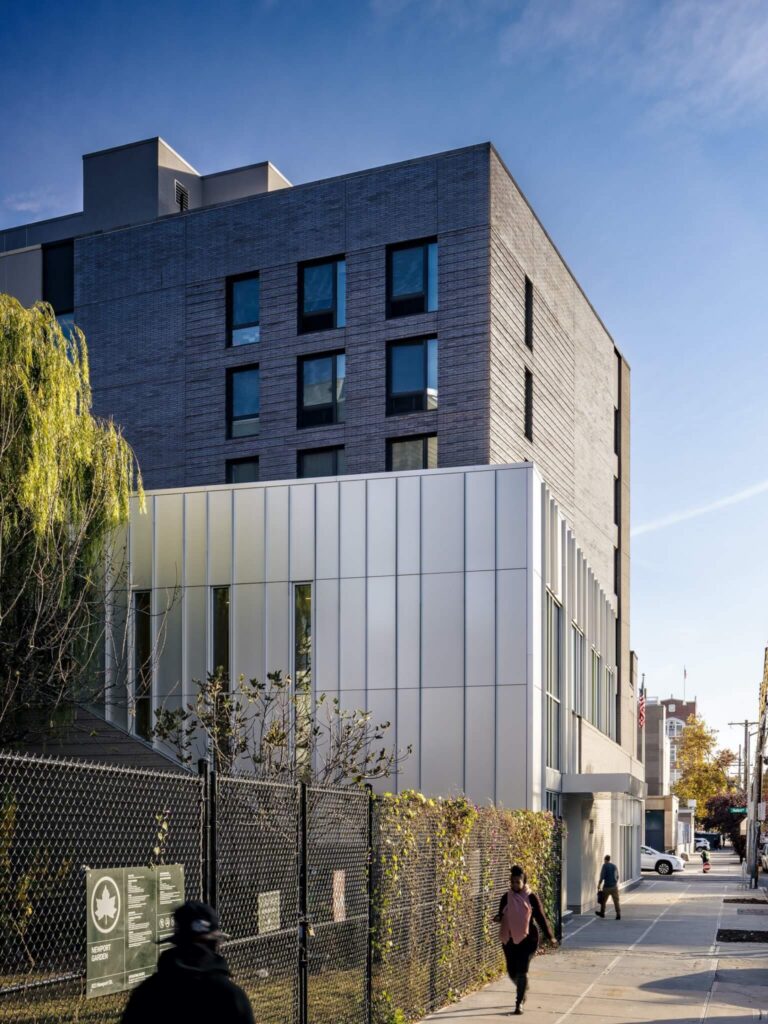Building exterior in sunlight, showing the dark brick residential mass above a gleaming metallic panel block, emphasizing the functional separation in the Vertical Integration of Industry and Housing.