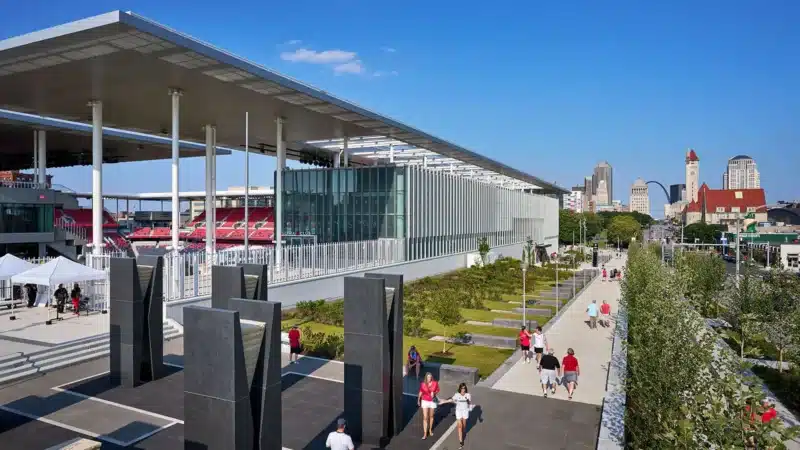 The stadium's exterior plaza overlooks the city skyline, featuring green landscapes and a sculptural monument documenting the site's history.