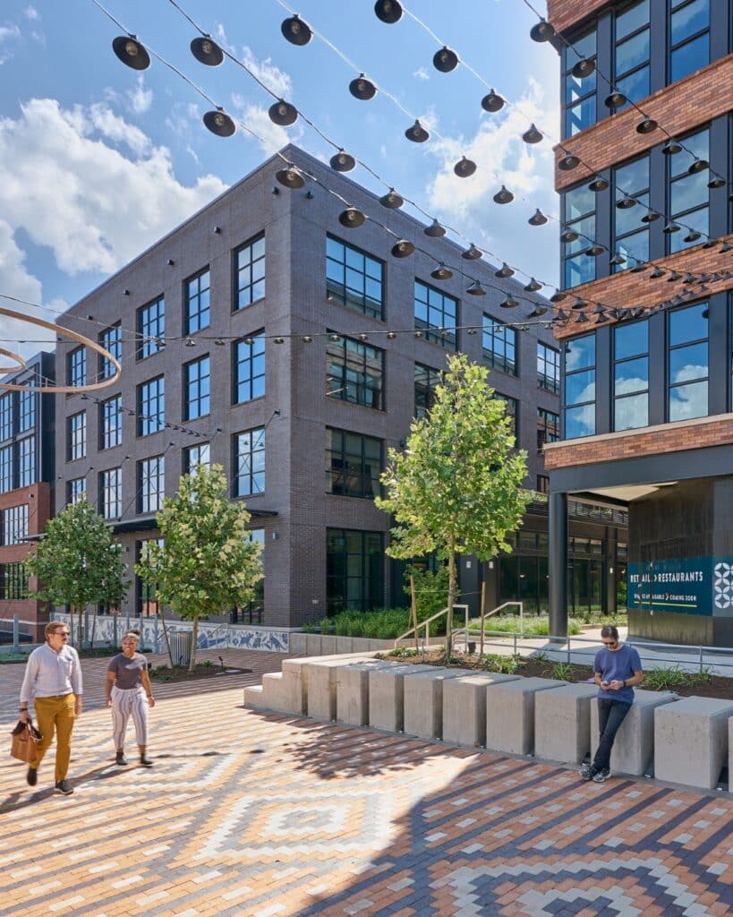 Pedestrians stroll through the public plaza at Centro West in Austin, surrounded by five-story brick buildings with black-framed windows and overhead string lights against a blue sky.