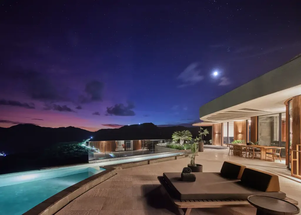 منظر ليلي لشرفة عقار نايرينستون في لومبوك مع مسبح لا نهائي، مقاعد خارجية، وسماء مرصعة بالنجوم فوق الخليج.