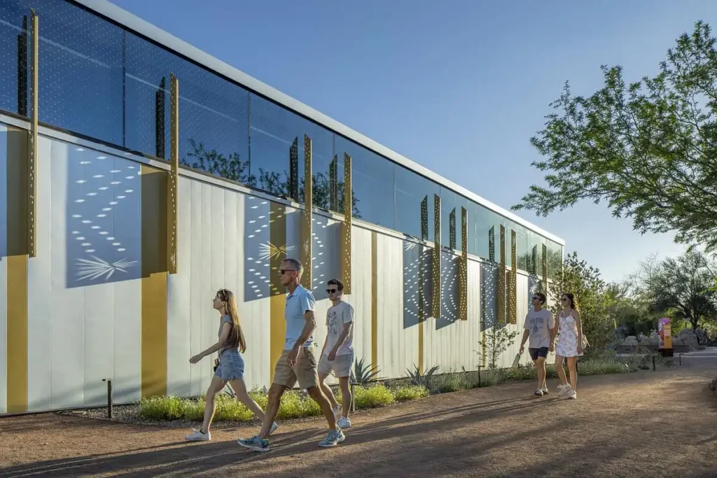 Visitors walk past the RAF Exhibit Building for Plant Science Phase II in Phoenix, featuring a glass facade with perforated metal screens casting dynamic shadows on white vertical panels.