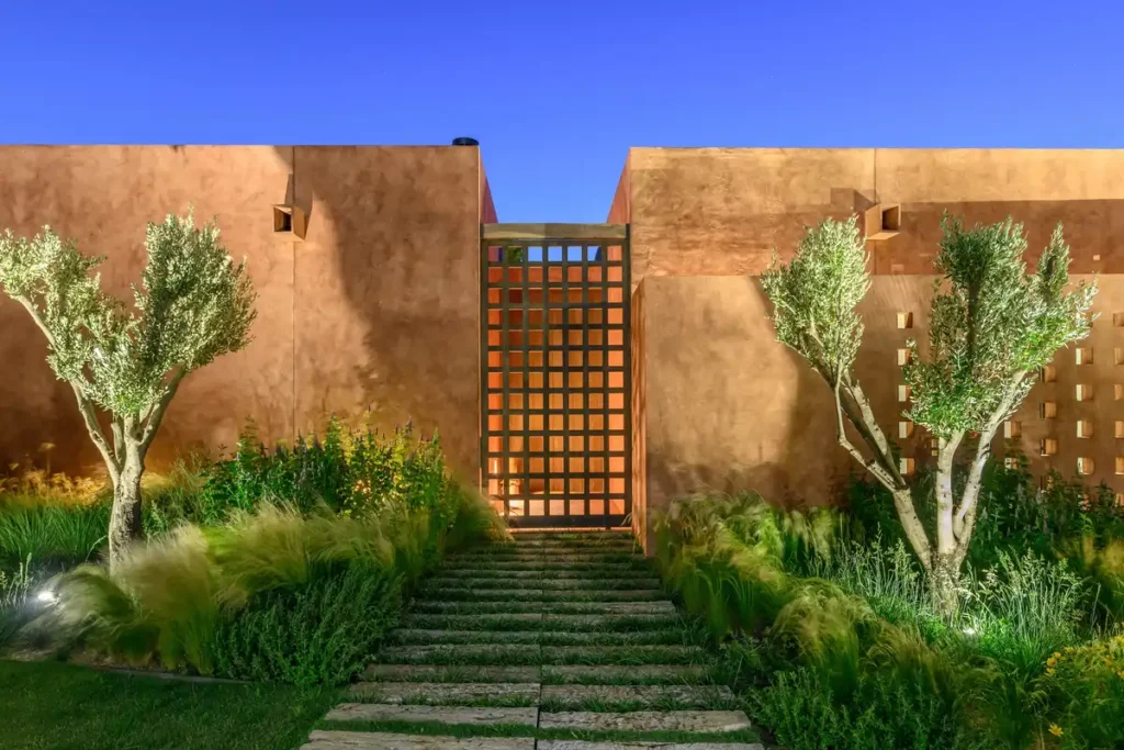 Stone steps rise through grasses to glowing lattice gate, earthy wall guards privacy.