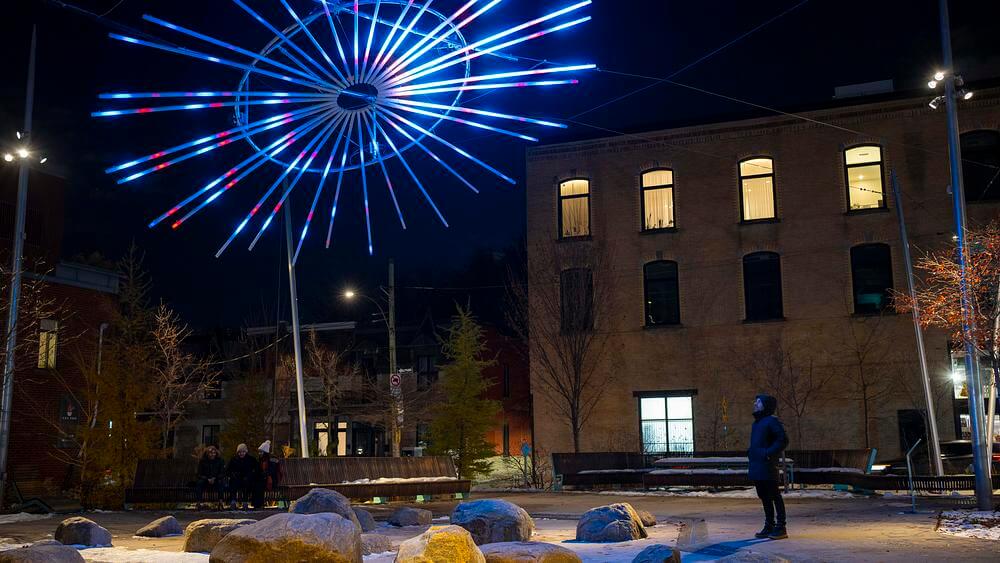 Un Brin d&rsquo;Soleil light installation glowing in blue and purple hues above Place des Fleurs-de-Macadam at night, with snow-covered ground and surrounding brick buildings.