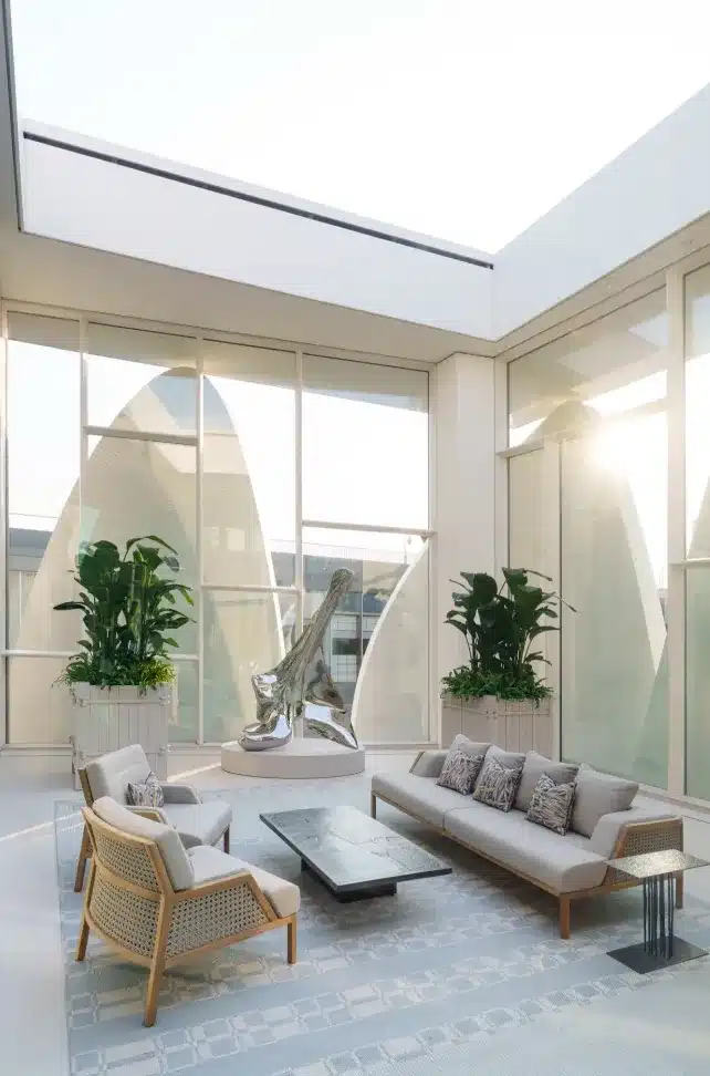 A sunlit interior lounge at the Dior Beijing flagship store, featuring modern furniture, large potted plants, and a reflective sculpture under a skylight.