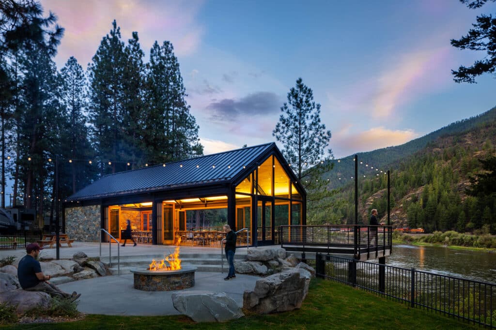 River Edge Pavilion at dusk with illuminated interior, outdoor fire pit, and guests enjoying the riverfront setting.
