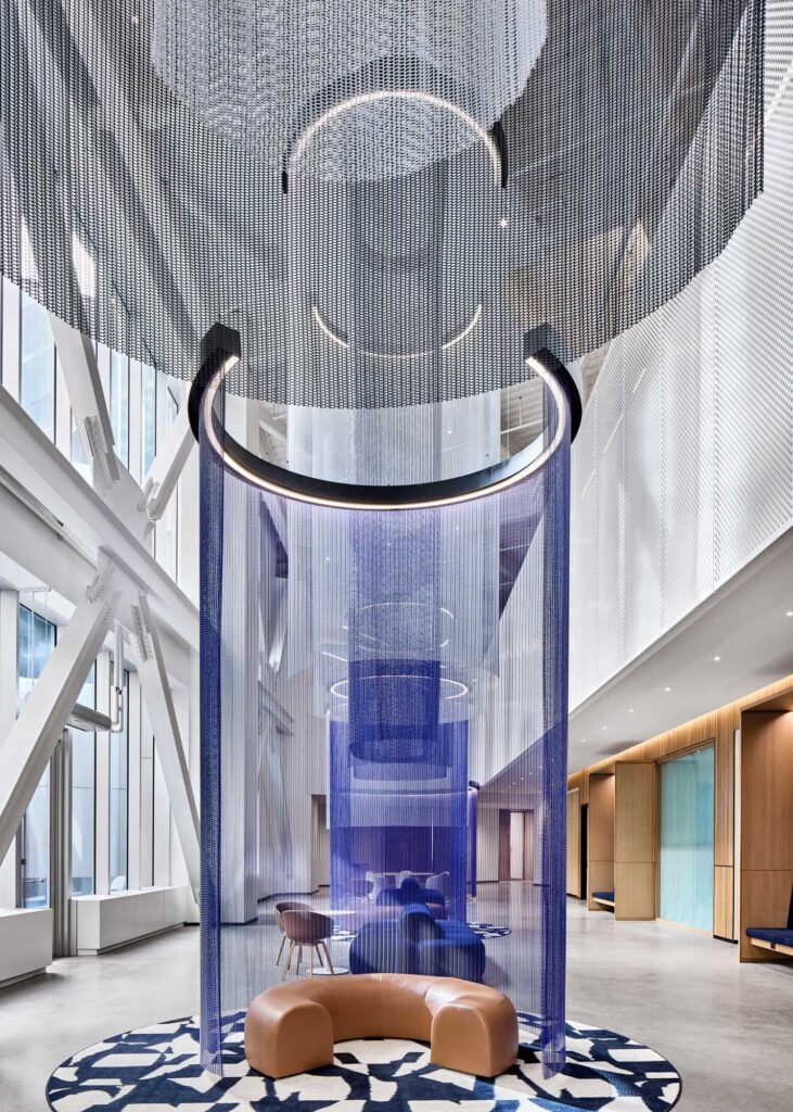 رواق داخلي مذهل في الحرم المركزي لشركة فورد، يضم تمثالًا معلقًا من الشبكة المعدنية الزرقاء مع إضاءة دائرية مدمجة، يحيط بمنطقة جلوس من الجلد المنحني على سجادة بنمط هندسي، تحت سقف عالٍ مع عوارض هيكلية بيضاء ظاهرة.