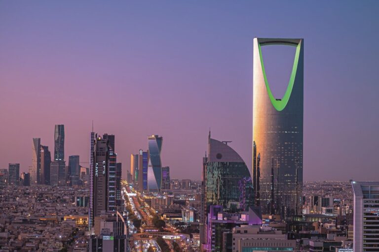 A panoramic evening view of the Riyadh skyline featuring the iconic Kingdom Centre tower illuminated in green against a purple sunset sky.