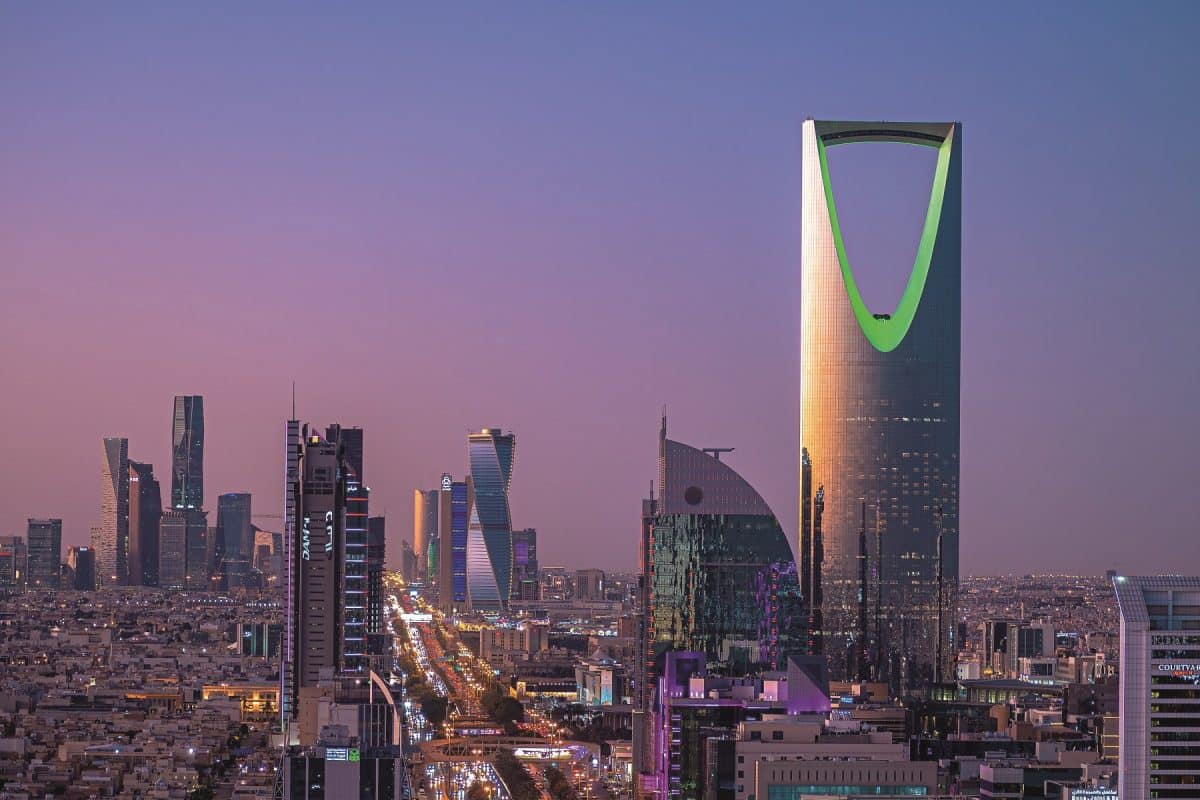 A panoramic evening view of the Riyadh skyline featuring the iconic Kingdom Centre tower illuminated in green against a purple sunset sky.