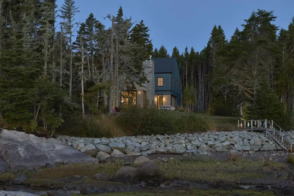 Warm light glows from windows, illuminating a stone entry nestled between dense forest and coastal rocks.