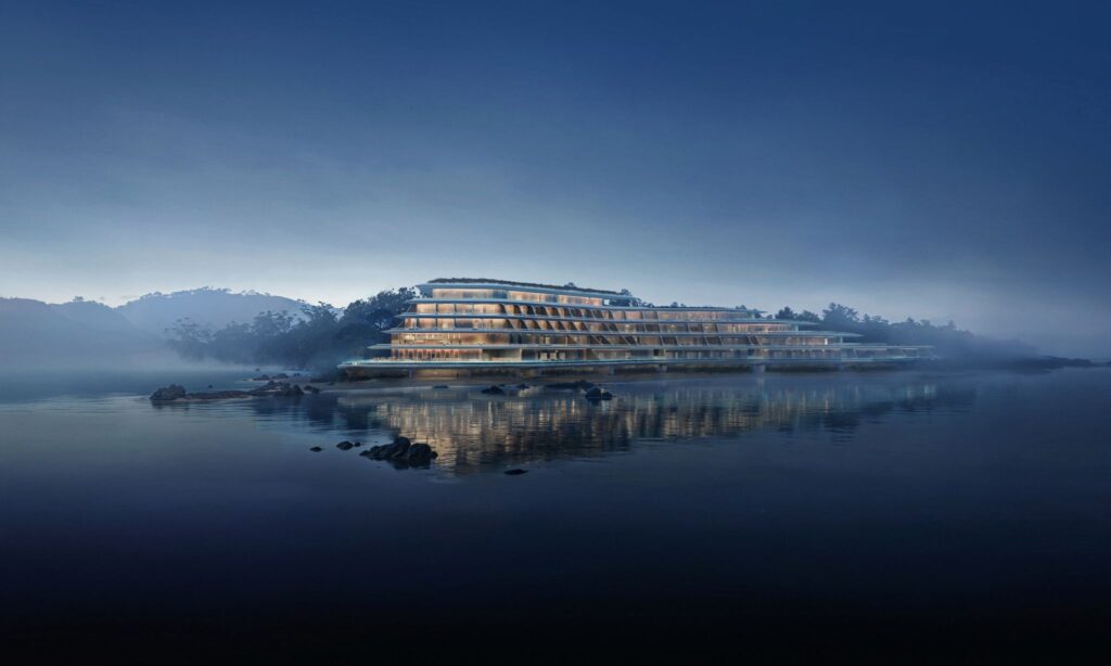 NOT A HOTEL Okinawa illuminated at dusk, its terraced structure reflecting on calm waters, nestled between forest and sea.