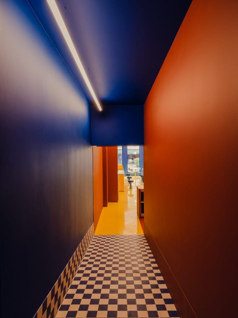 A narrow blue-and-orange corridor leads to light, checkered floor emphasizing directional flow.