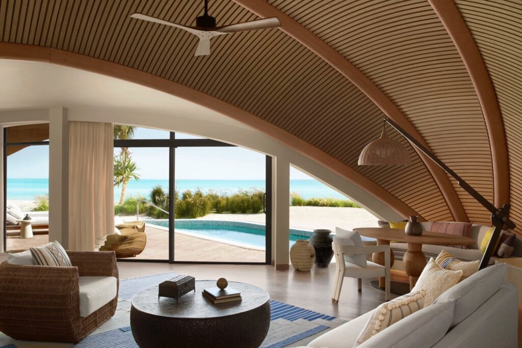 Luxury villa interior with curved wooden ceiling, woven furniture, and floor-to-ceiling windows overlooking a private pool and ocean view.