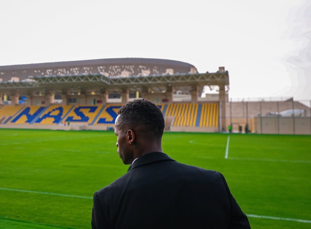 A man in formal attire stands on Al-Nassr&rsquo;s training pitch, gazing toward the stadium with &ldquo;NASSR&rdquo; lettering visible in blue and yellow seats.