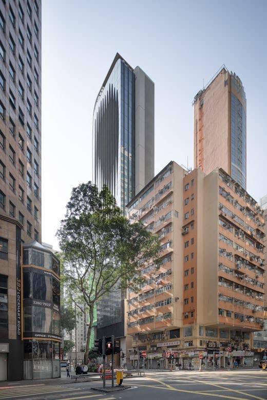 منظر للشارع في هونغ كونغ يظهر برج مكتب حديث بواجهة زجاجية بين مباني سكنية تقليدية، مع شجرة تضفي لمسة طبيعية على المشهد الحضري.