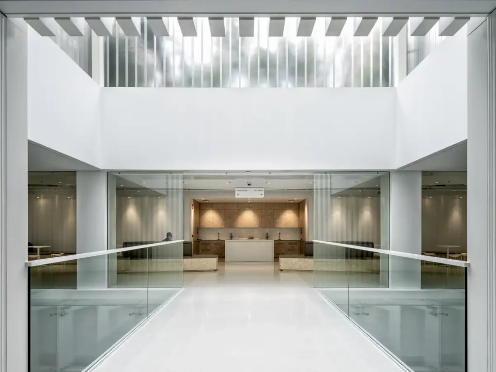 An open breakout area viewed from a glass walkway, featuring wooden cabinetry and seating in the background, highlighting the balance between transparency and natural materials in the Sustainable Headquarters Design.