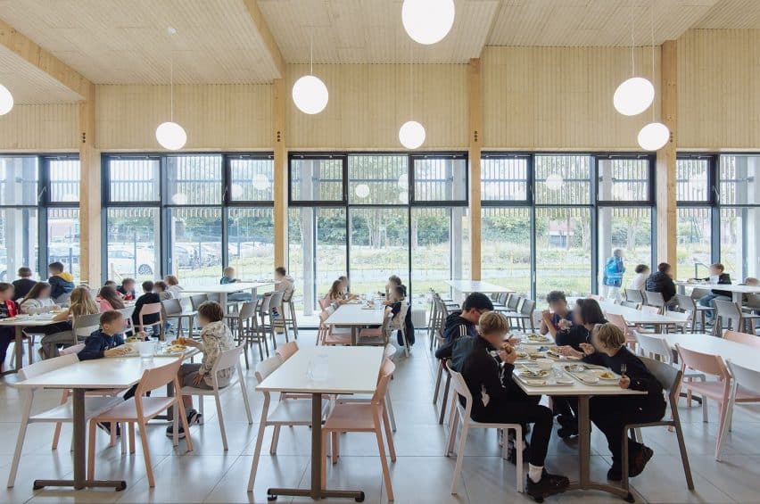 Canteen of the timber-framed school in Cambrai, showing exposed wooden columns, a spacious floor, and students eating under natural and artificial lighting.