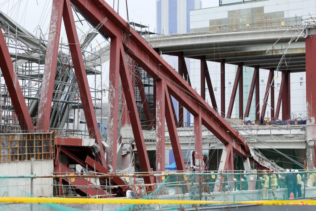 Scene from the Gwangju library collapse site, showing red steel trusses collapsed over shattered concrete slabs between floors, with rescue teams inspecting debris after a concrete pouring accident that killed four workers.
