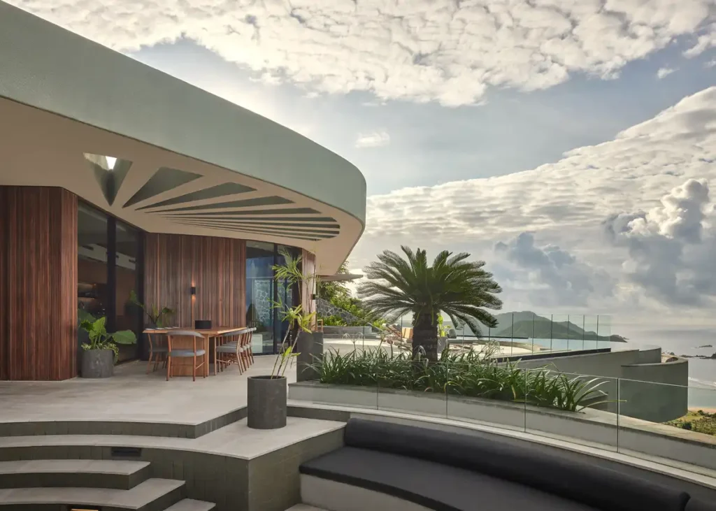 شرفة عقار ذات سقف منحني، وواجهة خشبية من الساج، وسياج زجاجي يطل على البحر تحت سماء ملبدة بالغيوم.