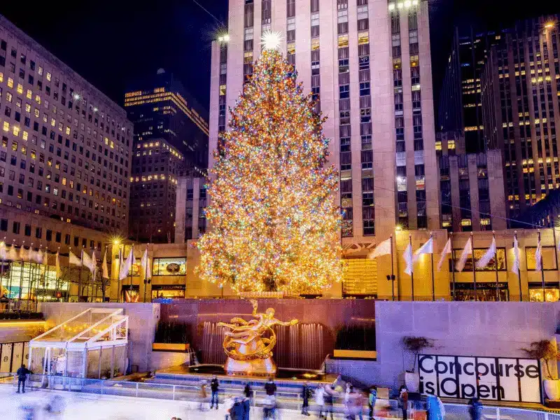 The Rockefeller Center Christmas tree glows with 50,000+ LED lights at night, crowned by a radiant star, with skaters on the ice rink below and Art Deco buildings framing the scene.