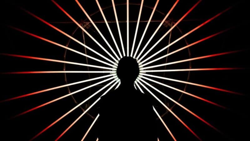 Silhouette of a person standing beneath the glowing radial structure of Un Brin d&rsquo;Soleil at night, with alternating red and white light beams radiating outward against a dark sky.