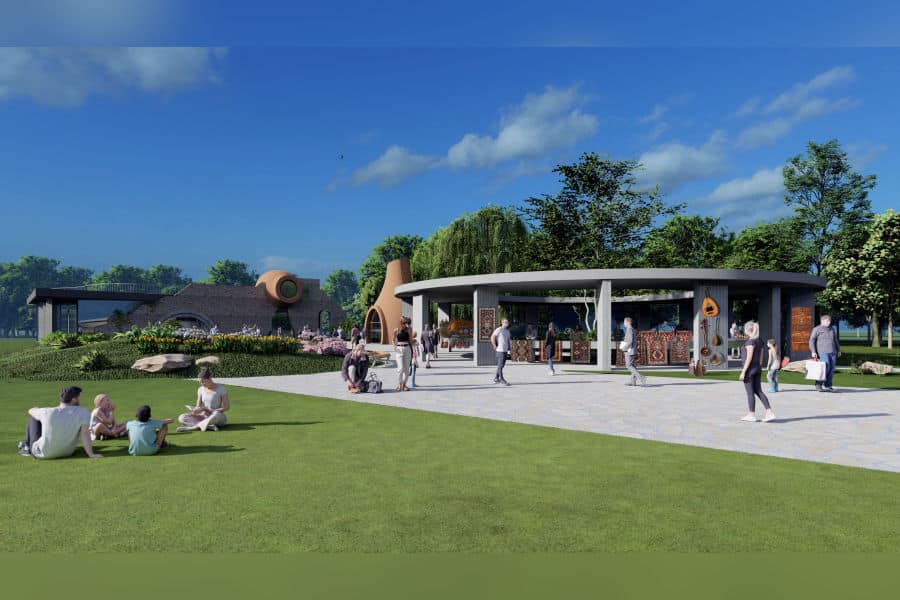 يوم مشمس في حديقة أرتا، يُظهر الزوار وهم يستريحون على العشب ويسيرون قرب القاعة الدائرية للسوق، مع هياكل طينية رمزية ونباتات خضراء تحت سماء زرقاء.