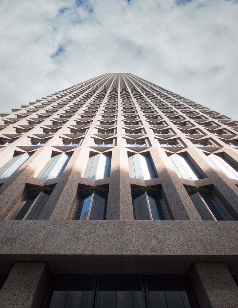 مشهد من الأسفل لبرج خرساني عالٍ يحمل نمطًا هندسيًّا متكررًا للنوافذ وشرفات بارزة، أمام سماء جزئيًا غائمة.