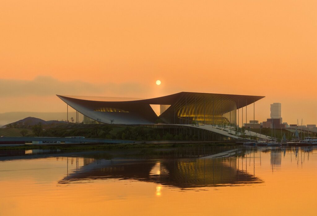 Ulsan Performing Arts Venue at sunset, its sweeping roof and glass facade reflected in the river. The  bridges urban and natural landscapes.