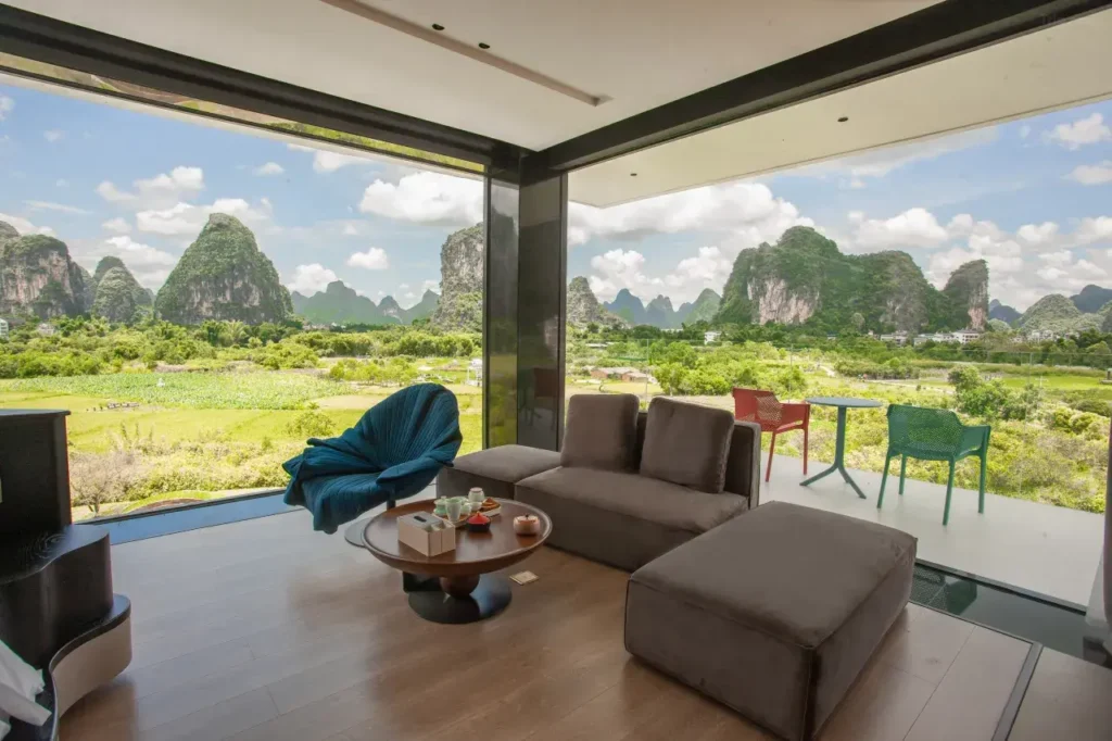 Modern hotel lounge with floor-to-ceiling windows overlooking lush green fields and iconic karst mountains in Guilin, featuring minimalist furniture and a glass balcony. Avatar like views