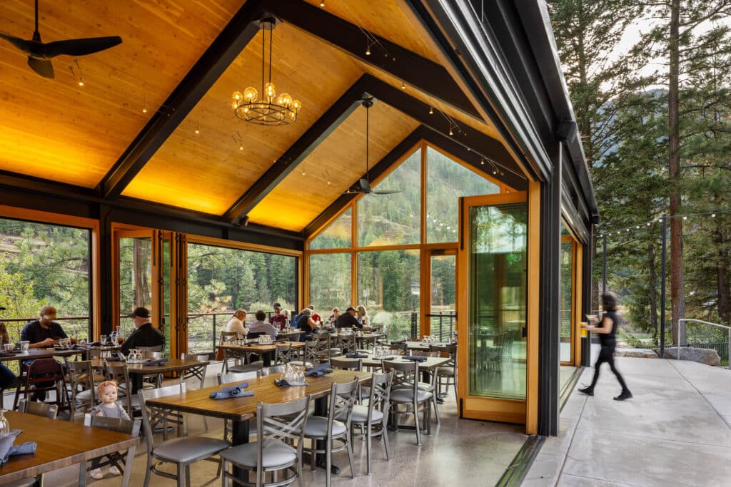 Dining area inside River Edge Pavilion with guests seated at tables under a vaulted wood ceiling and large glass windows overlooking forested hills.
