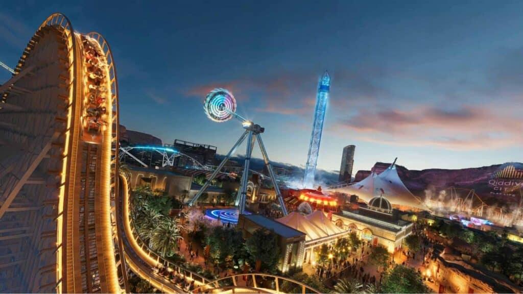 A vibrant evening rendering of Six Flags Qiddiya City featuring illuminated roller coasters, a giant swinging pendulum ride, and a drop tower against a twilight desert sky.