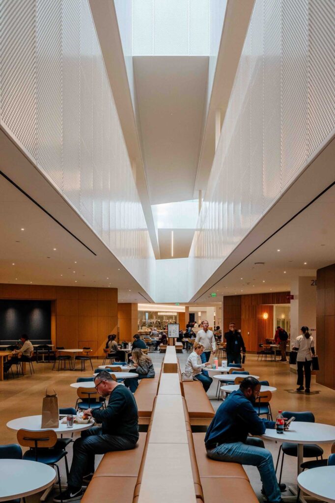 Employees dining and working in Ford&rsquo;s Central Campus food hall, featuring long communal benches, round tables, and a soaring ceiling with perforated white panels that diffuse natural light from skylights above.