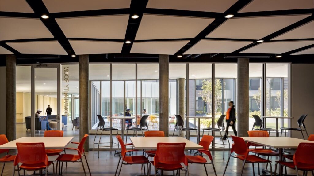 Well-lit indoor community space, used for dining and meetings, overlooking the exterior courtyard through a glass wall, enhancing the Vertical Integration of Industry and Housing on a social level.