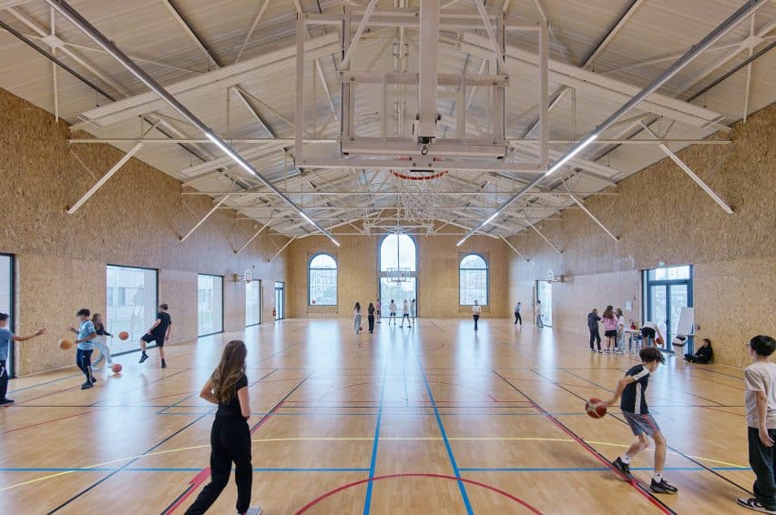 Interior gym of the timber-framed school in Cambrai, showing OSB-clad walls, wooden flooring, and students playing basketball under an exposed structural ceiling.