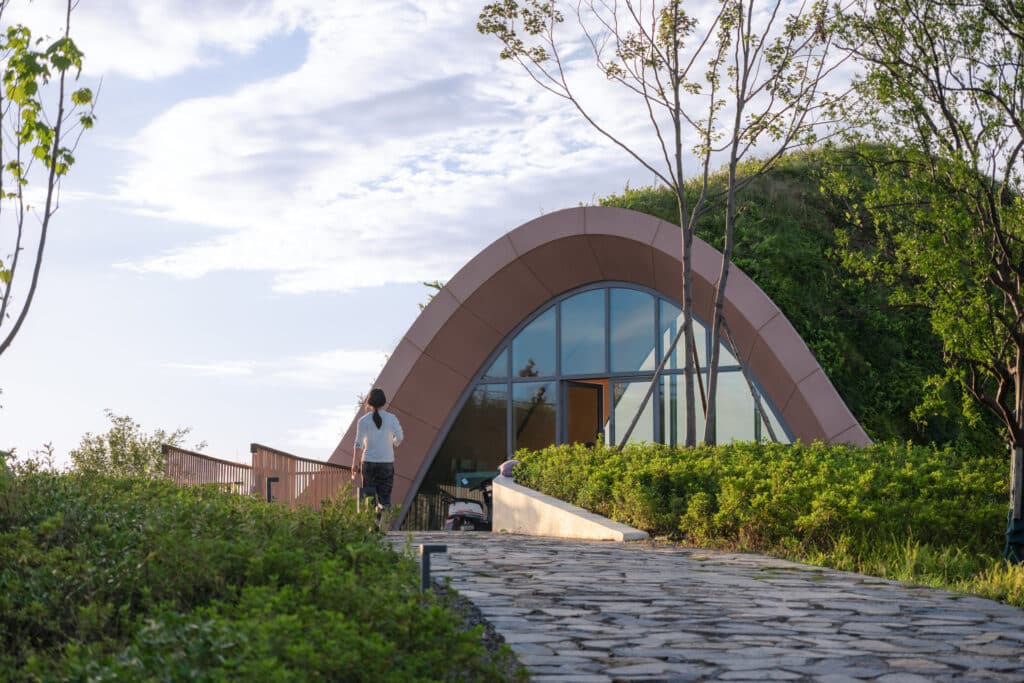 Pujiang Platform in Chengdu, a timber arched entrance blending into the green hill, with a visitor walking along a stone path toward the wide glass facade.