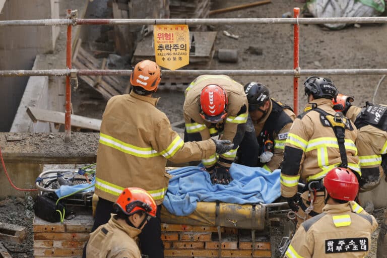South Korean firefighters attend to a victim at the Gwangju library collapse site, with a falling hazard warning sign visible amid concrete debris and metal structures.