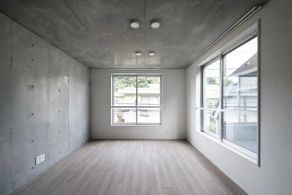 Empty interior room at Escenario Hanabusayama in Tokyo, featuring exposed concrete walls, light wood flooring, and dual-sided windows for natural ventilation.