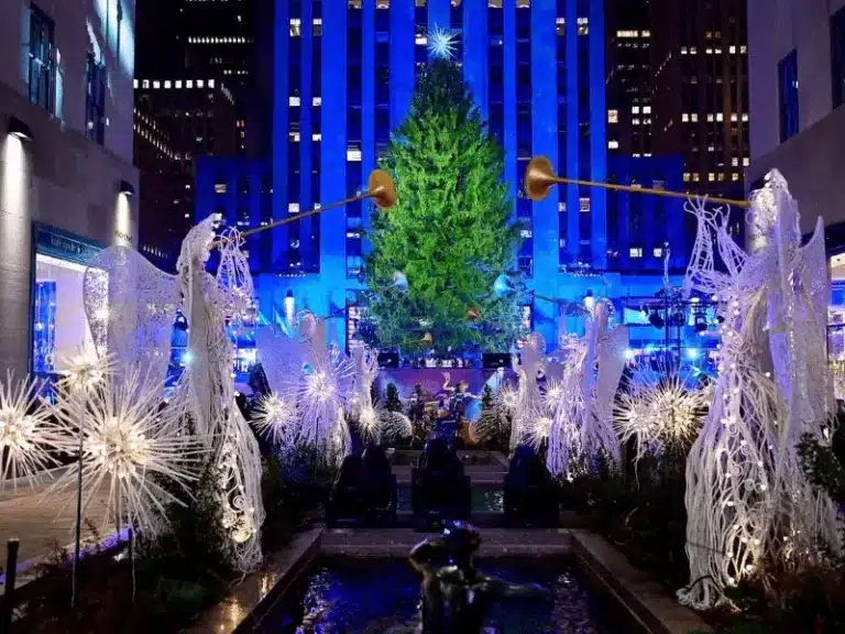 Illuminated angel sculptures with glowing trumpets surround the Rockefeller Center Christmas tree at night, set against a blue-lit Art Deco building facade.