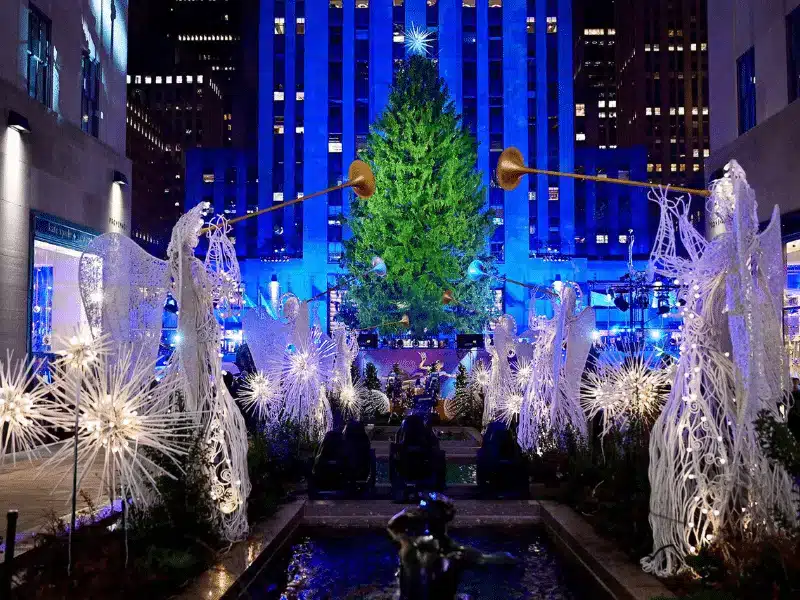 Illuminated angel sculptures with glowing trumpets surround the Rockefeller Center Christmas tree at night, set against a blue-lit Art Deco building facade.